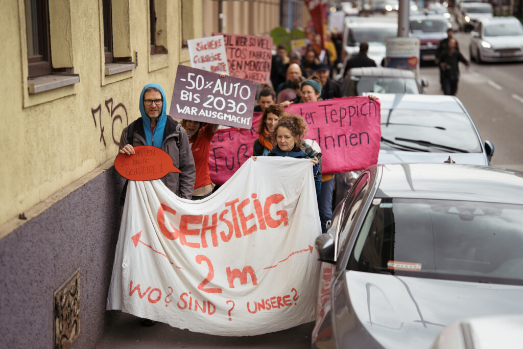 Auf diesem Foto der Aktion #WorstOfGÜRTELGehsteig ist ein Umzug an Demonstrierenden zu sehen, der zwischen Häuserwand und Gehsteigparkern entlang vom Inneren Neubaugürtel geht. Ganz vorne tragen zwei Personen einen großen Banner mit der Aufschrift "Gehsteige 2m?"
Foto Copyright (c) Tom Poe Photography