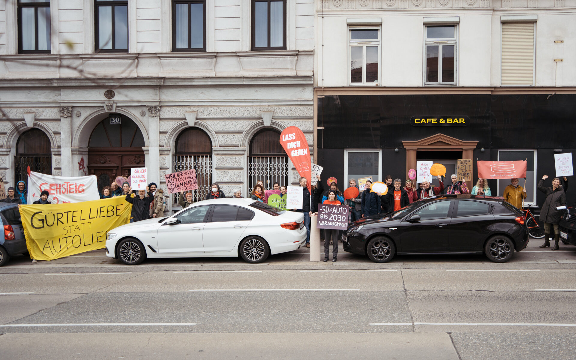 Das Bild zeigt im Vordergrund die vier leeren Gürtel-Fahrspuren für Kfz in eine Richtung, dann eine weitere Spur für Gehsteigparker. Hinter den parkenden Autos sind die Demo-Teilnehmenden mit bunten Schildern und Banndern zu sehen. Photo credits (c): Tom Poe Photography